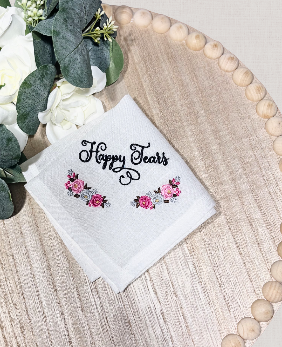 White handkerchief with 'Happy Tears' embroidered text and floral designs on a wooden surface with flowers.