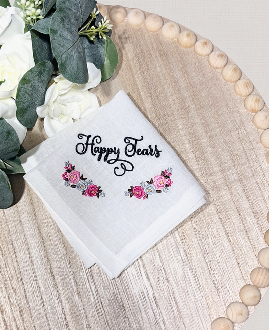 White handkerchief with 'Happy Tears' embroidered text and floral designs on a wooden surface with flowers.