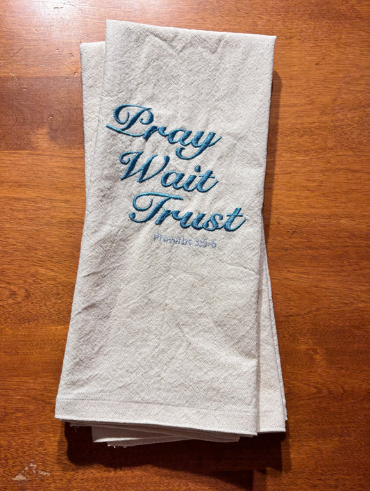 Folded towel with 'Pray Wait Trust' text on a wooden surface