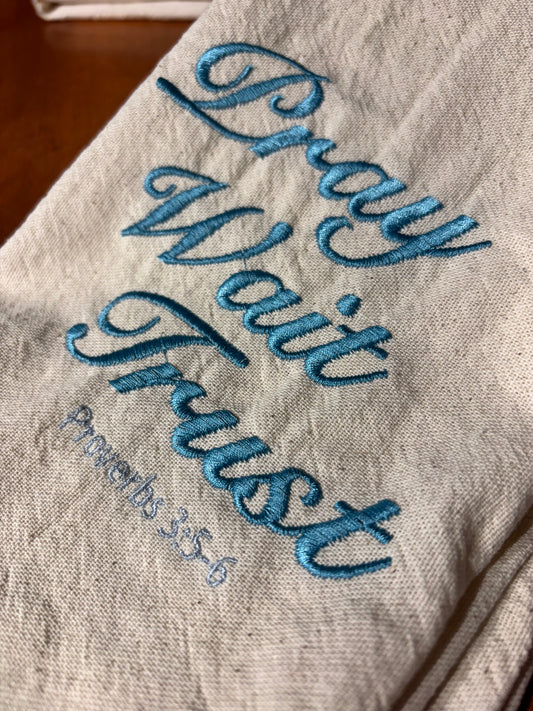 Textile with blue embroidered text 'Pray Wait Trust' on a wooden surface